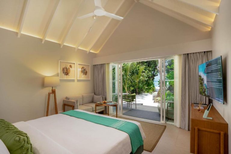 Interior de una Beach Villa en RAAYA by Atmosphere, Maldivas, con dormitorio elegante y vistas a la playa y la laguna turquesa al fondo.